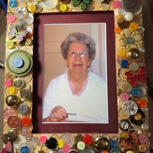 May include: A rectangular photo frame decorated with a variety of colorful buttons and butterfly embellishments. The frame surrounds a portrait of an elderly person. The buttons are in various sizes, colors, and materials, creating a unique, handcrafted look.