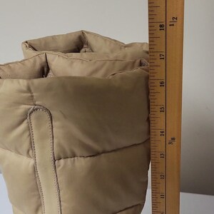 Pu&ograve; includere: Un paio di stivali invernali imbottiti beige con un dettaglio in pelle. Gli stivali sono misurati con un righello, mostrando che sono alti circa 43 cm.