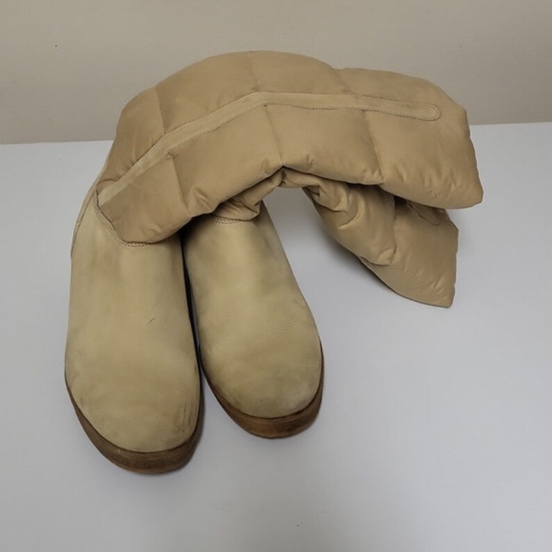 Pu&ograve; includere: Un paio di stivali alti al ginocchio in pelle scamosciata color beige con un design trapuntato a soffietto sulla parte superiore dello stivale.