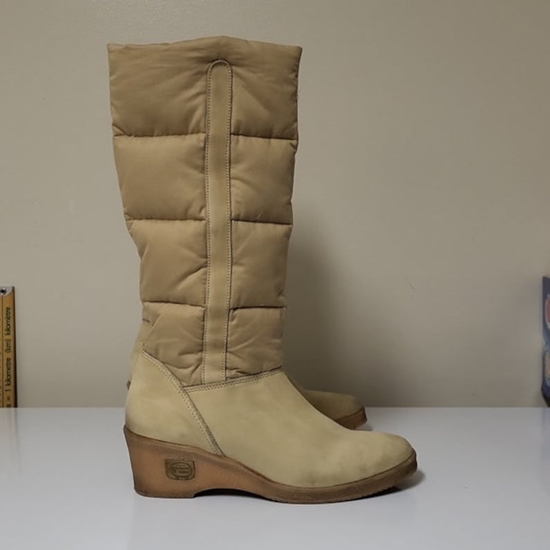 Pu&ograve; includere: Stivali beige imbottiti alti al ginocchio con tacco a zeppa. Gli stivali hanno una cerniera sul retro e una suola in pelle marrone.