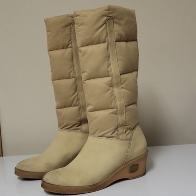 Pu&ograve; includere: Un paio di stivali beige alti al ginocchio con tacco a zeppa. Gli stivali sono realizzati in materiale trapuntato e hanno una cerniera sul retro.