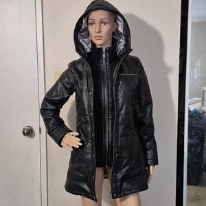 Puede incluir: Chaqueta negra de imitación de cuero con capucha, cierre de cremallera y diseño acolchado. La chaqueta tiene un cuello alto y una longitud larga.