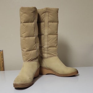 Pu&ograve; includere: Un paio di stivali beige alti al ginocchio con tacco a zeppa. Gli stivali sono realizzati in materiale trapuntato e hanno una finitura simile alla pelle scamosciata.