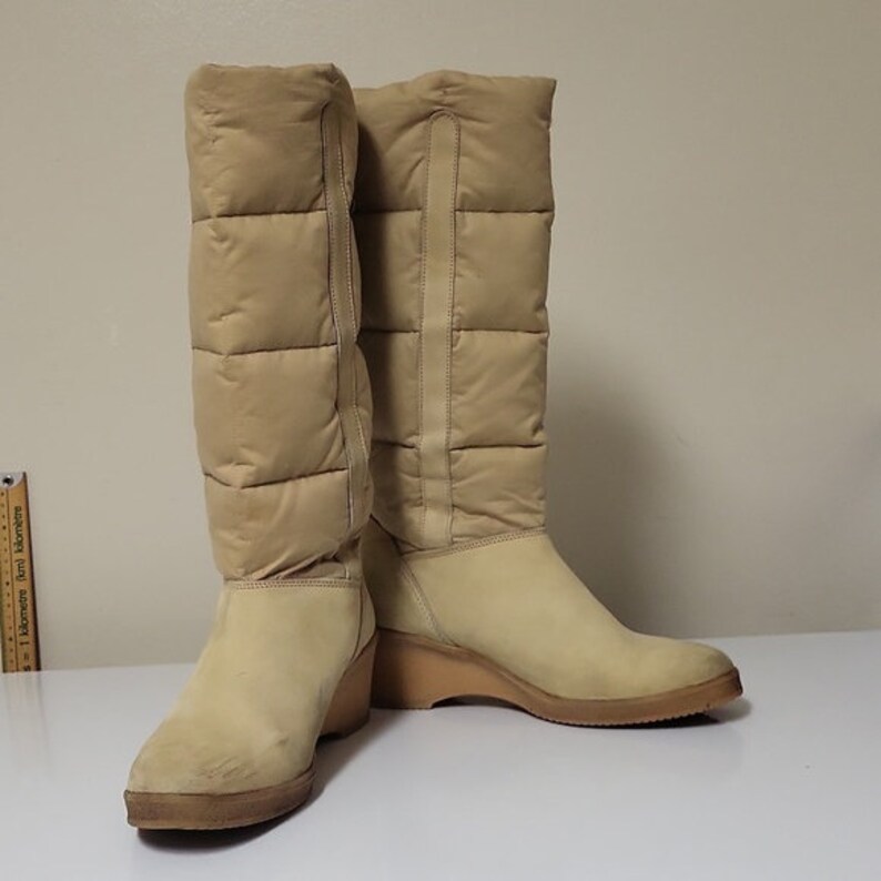 Pu&ograve; includere: Un paio di stivali beige alti al ginocchio con tacco a zeppa. Gli stivali sono realizzati in materiale trapuntato e hanno una finitura simile alla pelle scamosciata.