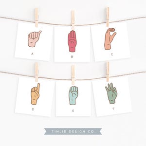 May include: Six cards with different hand gestures in American Sign Language. Each card has a letter of the alphabet (A-F) printed on it. The cards are hung on a string with clothespins.