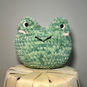 May include: A handmade, plush frog with a round body and a textured, green surface. The frog has black eyes, a black stitched mouth, and pink cheeks. The frog is sitting on a woven basket.
