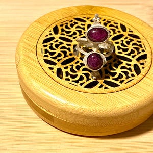 May include: A silver ring with two oval, deep red gemstones. The ring is set on a wooden box with a decorative, openwork design. The box and the surface it rests on are made of light-colored wood.