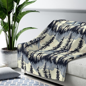May include: A soft, cream-colored throw blanket with a dark blue and light blue forest silhouette pattern. The blanket is draped over a white couch.