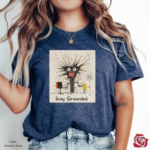 May include: A heather navy blue t-shirt with a graphic of a black cartoon character with red eyes and a yellow and orange background. The text "Stay Grounded" is printed below the graphic.
