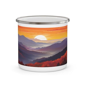 May include: White enamel camping mug with a colorful illustration of a mountain range at sunset. The mountains are purple, blue, and green, with a bright orange sky and a large yellow sun.