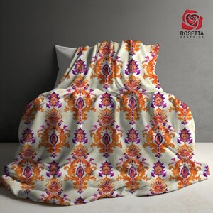 May include: A white throw blanket with a repeating pattern of orange and purple floral designs. The blanket is folded and draped over a pillow.
