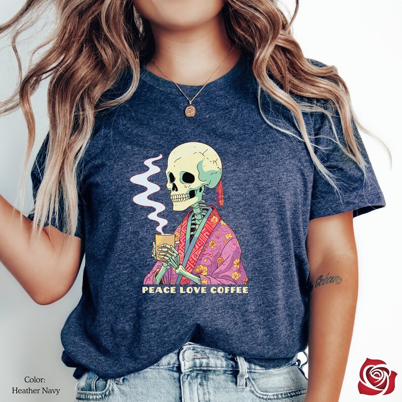 May include: A heather navy blue t-shirt featuring a skeleton in a pink and floral robe holding a cup of coffee with steam rising from it. The text "Peace Love Coffee" is printed below the skeleton.