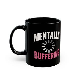 Puede incluir: Taza de cerámica negra con las palabras "MENTALLY BUFFERING" en blanco y rosa. El diseño incluye un símbolo de carga. La taza tiene un asa curva.