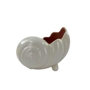 May include: A ceramic seashell-shaped planter in a pearlescent white finish. The interior is a deep reddish-brown. The planter has a spiral design and rests on three small, rounded feet. A decorative item for coastal or nautical themes.