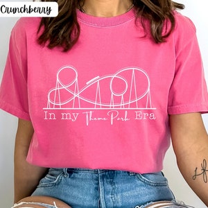 May include: A pink t-shirt with a white outline of a roller coaster and the text "In my Theme Park Era".