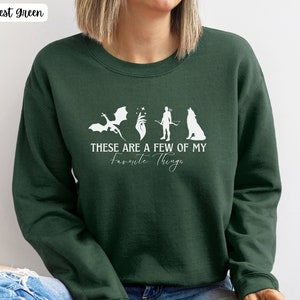 May include: Forest green sweatshirt with a white graphic of a dragon, a hand with a magic wand, a person with a bow and arrow, and a wolf. The text on the sweatshirt reads "These are a few of my favorite things."