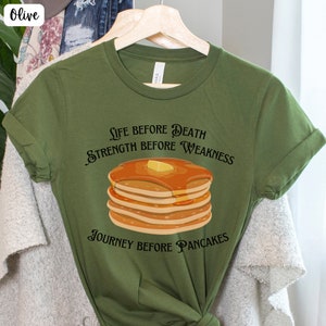 May include: Olive green t-shirt with a graphic of a stack of pancakes and the text "Life before death, strength before weakness, journey before pancakes."