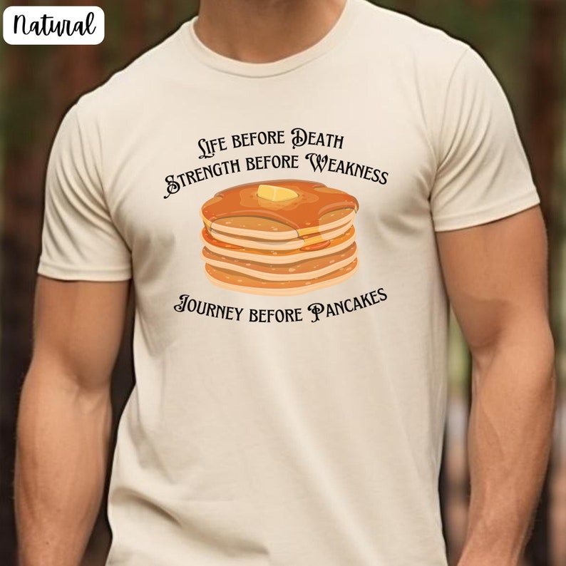 May include: A beige t-shirt with a graphic of a stack of pancakes and the text "Life before Death, Strength before Weakness, Journey before Pancakes".