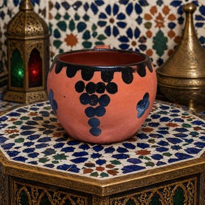 Peut inclure: Un bol en céramique de couleur terre cuite avec un bord noir et un motif de grappe de raisin. Le bol est décoré d'accents bleus et repose sur une table carrelée en mosaïque. Des lanternes décoratives et un récipient en laiton sont en arrière-plan.