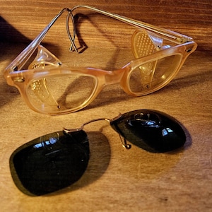 May include: Vintage eyeglasses with a light brown frame and clear lenses, featuring side shields. A separate pair of black clip-on sunglasses rests below. The glasses have a classic, retro design.