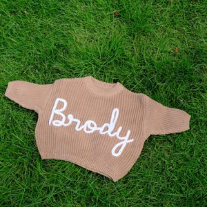 K&ouml;nnte beinhalten: Ein beigefarbener, lang&auml;rmeliger Strickpullover mit Rundhalsausschnitt und gerippter Textur. Das Wort Brody ist in wei&szlig;er Schreibschrift mittig auf der Brust aufgestickt. Das Kleidungsst&uuml;ck ist in einem dicken, groben Strickmuster gefertigt.