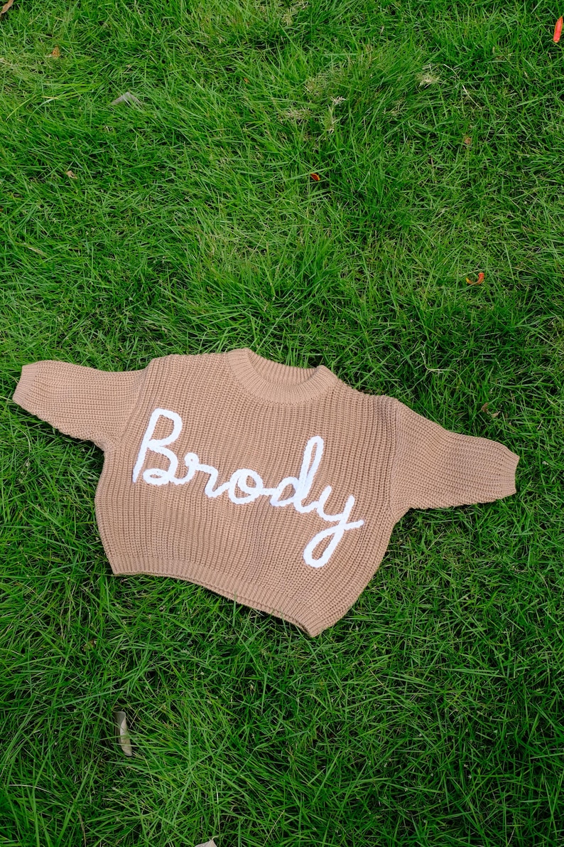 K&ouml;nnte beinhalten: Ein beigefarbener, lang&auml;rmeliger Strickpullover mit Rundhalsausschnitt und gerippter Textur. Das Wort Brody ist in wei&szlig;er Schreibschrift mittig auf der Brust aufgestickt. Das Kleidungsst&uuml;ck ist in einem dicken, groben Strickmuster gefertigt.