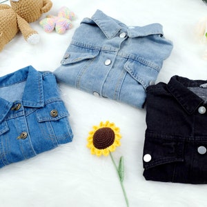 May include: Three denim jackets in different colors: light blue, dark blue, and black. The jackets are laid out on a white surface with a crocheted sunflower and a crocheted teddy bear.