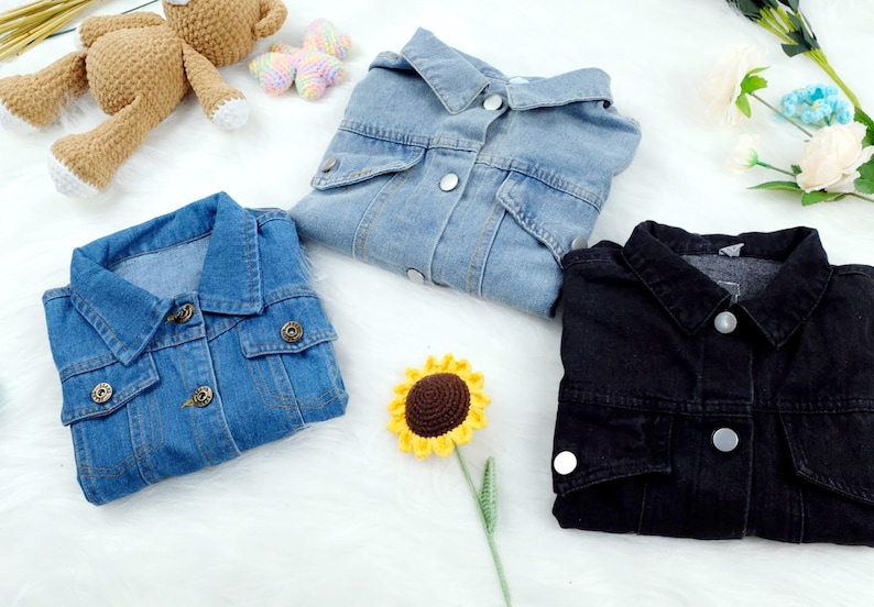 May include: Three denim jackets in different colors: light blue, dark blue, and black. The jackets are laid out on a white surface with a crocheted sunflower and a crocheted teddy bear.