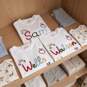 May include: Three white short-sleeved t-shirts featuring embroidered names Sam, Wells, and Watson in colorful yarn. Each shirt includes a small embroidered apple, a car, and the letters ABC. The shirts are displayed alongside folded striped and patterned clothing.