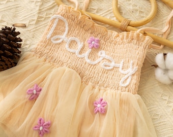 Puede incluir: Un body de bebé de color melocotón pálido con una falda de tul y la palabra "Daisy" bordada en blanco. El body tiene adornos de flores rosas.