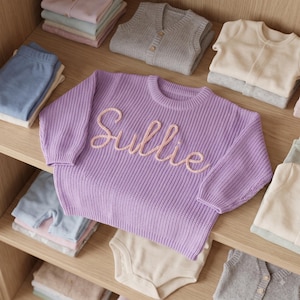 K&ouml;nnte beinhalten: Ein lang&auml;rmeliger Strickpullover in Lavendel mit dem in hellrosa Garn auf die Brust gestickten Namen Sullie. Der Pullover hat einen gerippten Rundhalsausschnitt, B&uuml;ndchen und Saum und liegt zwischen verschiedenen gefalteten Kleidungsst&uuml;cken auf Holzregalen.