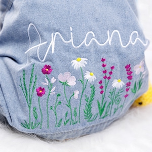 Puede incluir: Una chaqueta vaquera azul claro con el nombre "Ariana" bordado en la espalda. La chaqueta presenta una fila de flores bordadas en tonos rosa, blanco y verde.