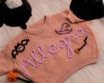 Può includere: Un body per neonati in maglia rosa chiaro con il nome "Allegra" in filato lavanda. Il body presenta un pipistrello nero, un cappello da strega e una scopa. Lo sfondo è una superficie bianca e soffice, con decorazioni sospese in bianco e nero.