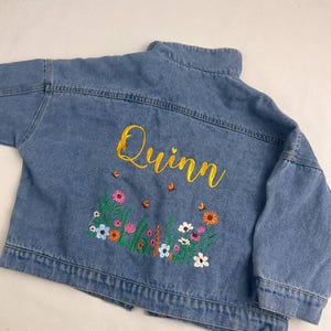 May include: A light blue denim jacket with the name "Quinn" embroidered in yellow script across the back. The jacket features a colorful floral embroidery design with pink, orange, and white flowers.