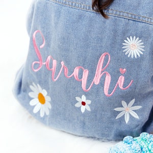 May include: A light blue denim jacket with pink embroidery that says "Sarahi" and white embroidered flowers.
