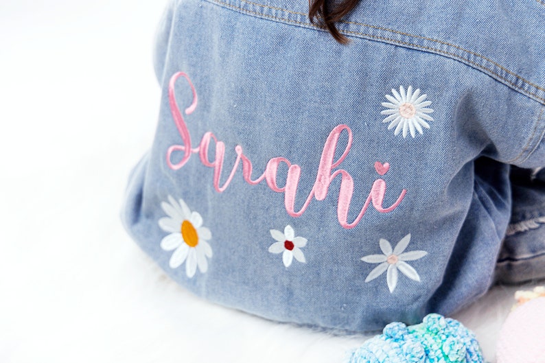 May include: A light blue denim jacket with pink embroidery that says "Sarahi" and white embroidered flowers.