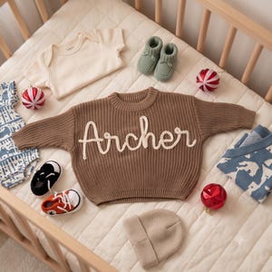 K&ouml;nnte beinhalten: Ein brauner Strickpullover mit dem in wei&szlig;er Schreibschrift gestickten Namen Archer, begleitet von einem cremefarbenen Body, salbeigr&uuml;nen Babyschuhen, einer beigen M&uuml;tze, einem blau gemusterten Body, einem blau-wei&szlig;en Pullover und zwei Paar Babyschuhen.