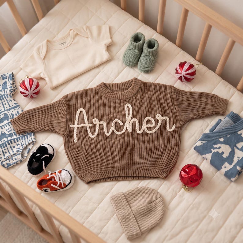 K&ouml;nnte beinhalten: Ein brauner Strickpullover mit dem in wei&szlig;er Schreibschrift gestickten Namen Archer, begleitet von einem cremefarbenen Body, salbeigr&uuml;nen Babyschuhen, einer beigen M&uuml;tze, einem blau gemusterten Body, einem blau-wei&szlig;en Pullover und zwei Paar Babyschuhen.