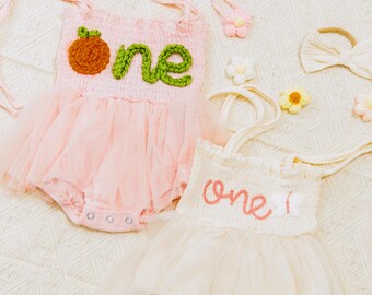 May include: Two pink and white tulle baby outfits with the word "one" embroidered on the front. The outfits are decorated with crocheted flowers and bows.