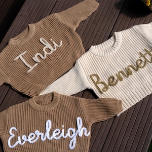 May include: Three personalized children's sweaters in shades of brown and cream. Each sweater features a name embroidered across the chest: "Indi", "Bennett", and "Everleigh". The sweaters are made of a ribbed knit material.