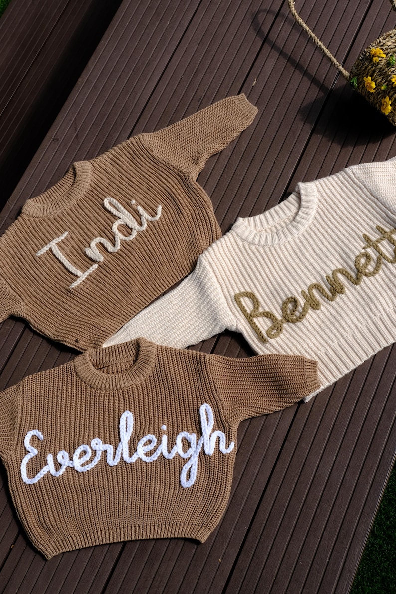 K&ouml;nnte beinhalten: Drei lang&auml;rmelige Strickpullover mit gerippter Textur und Rundhalsausschnitt. Zwei Pullover sind braun mit den wei&szlig; gestickten Namen 'Indi' und 'Everleigh', w&auml;hrend ein Pullover cremefarben mit dem braun gestickten Namen 'Bennett' ist.