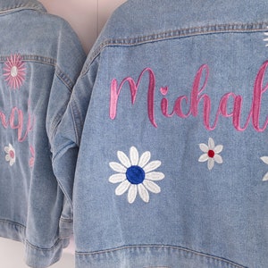 May include: Two denim jackets with embroidered flowers and names. The jacket on the left has the name "Sargis" embroidered in pink and the jacket on the right has the name "Michal" embroidered in pink.