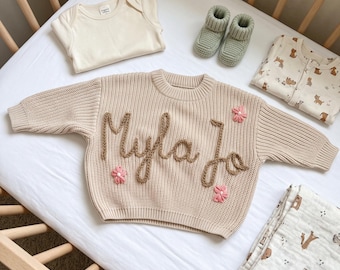 May include: A beige knit sweater featuring the embroidered name Myla Jo in brown yarn, accented with three small pink flowers. The set includes a plain cream bodysuit, a pair of sage green knit booties, and two pieces of patterned baby clothing.