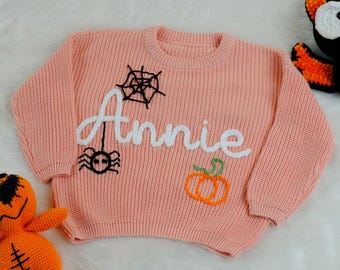 May include: A pink knitted sweater with a white embroidered name "Annie", a spider, a web, and a pumpkin.