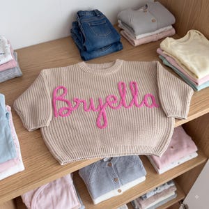 K&ouml;nnte beinhalten: Ein beigefarbener, lang&auml;rmeliger Strickpullover mit dem in leuchtend rosa Garn auf die Brust gestickten Namen Bryella. Der Pullover hat einen gerippten Rundhalsausschnitt, B&uuml;ndchen und Saum und liegt auf einem Holzregal zwischen gefalteter Kleidung.