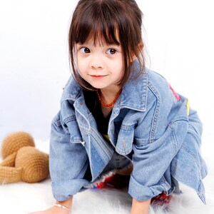May include: A young girl with brown hair wearing a blue denim jacket and a necklace. She is sitting on a white fluffy surface and looking at the camera.