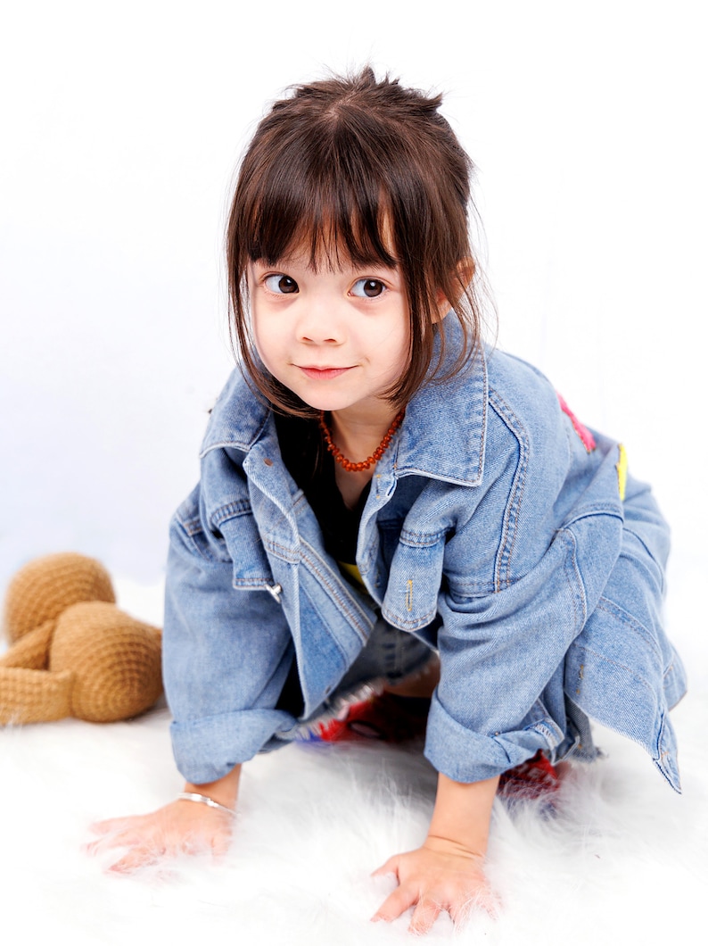 May include: A young girl with brown hair wearing a blue denim jacket and a necklace. She is sitting on a white fluffy surface and looking at the camera.