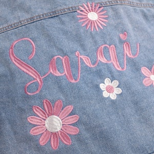May include: A light blue denim jacket with pink embroidery of the name "Sarai" and pink and white daisy flowers.