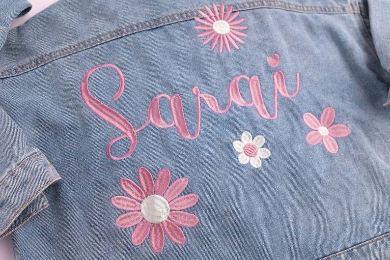 May include: A light blue denim jacket with pink embroidery of the name "Sarai" and pink and white daisy flowers.