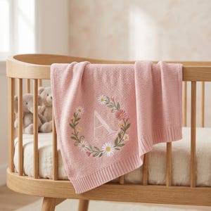 Könnte beinhalten: Eine rosa Babydecke mit einer floralen Kranzstickerei und dem Buchstaben "A". Die Decke ist über ein Holzbettchen drapiert. Der Kranz zeigt weiße Gänseblümchen, rosa Blüten und grüne Blätter.
